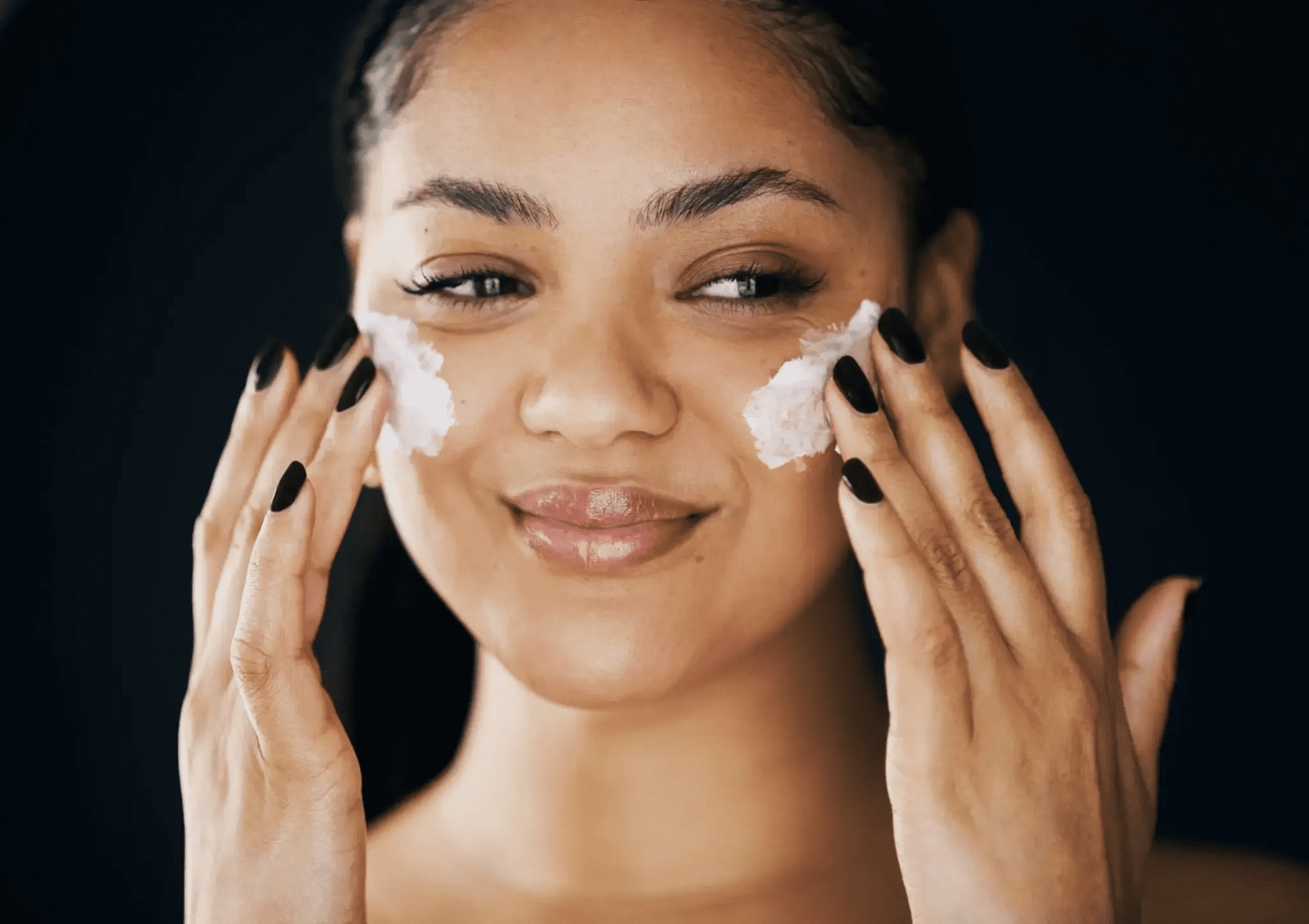 Woman applying moisturizer to her face with a smile.